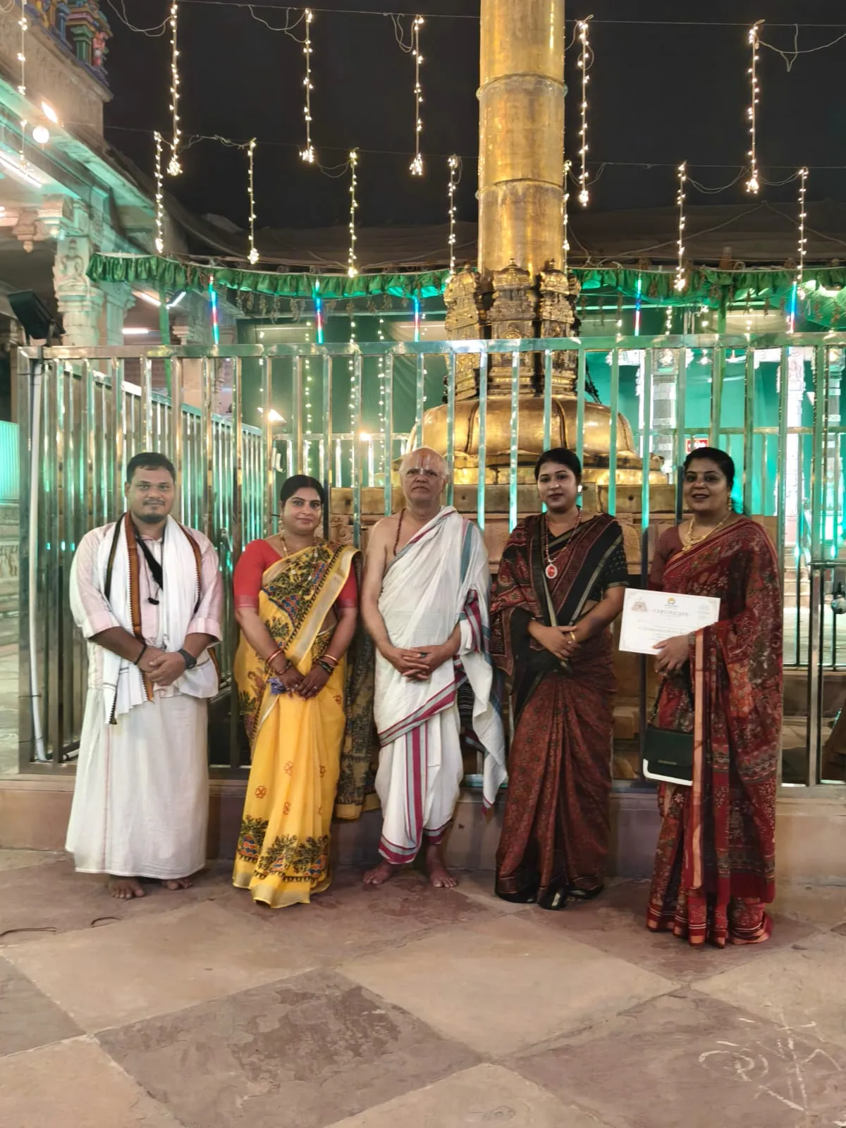 Certificate presentation beneath the golden pillar of an illuminated temple — Uddharsha Foundation cultural programme
