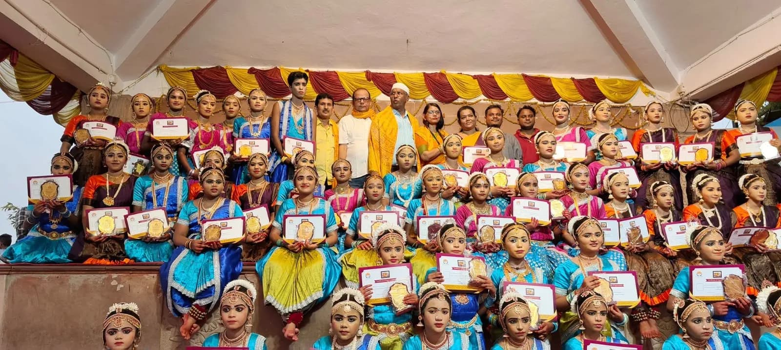 Large ensemble of young dancers holding certificates and awards on a decorated festival stage — Uddharsha Foundation